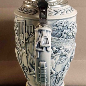 Puede incluir: Una jarra de cerveza de cer&aacute;mica con un dise&ntilde;o en relieve detallado que representa escenas ecuestres. La jarra tiene una tapa met&aacute;lica con bisagras, un asa decorativa y una banda colorida alrededor de la base. La combinaci&oacute;n de colores es blanca y azul.