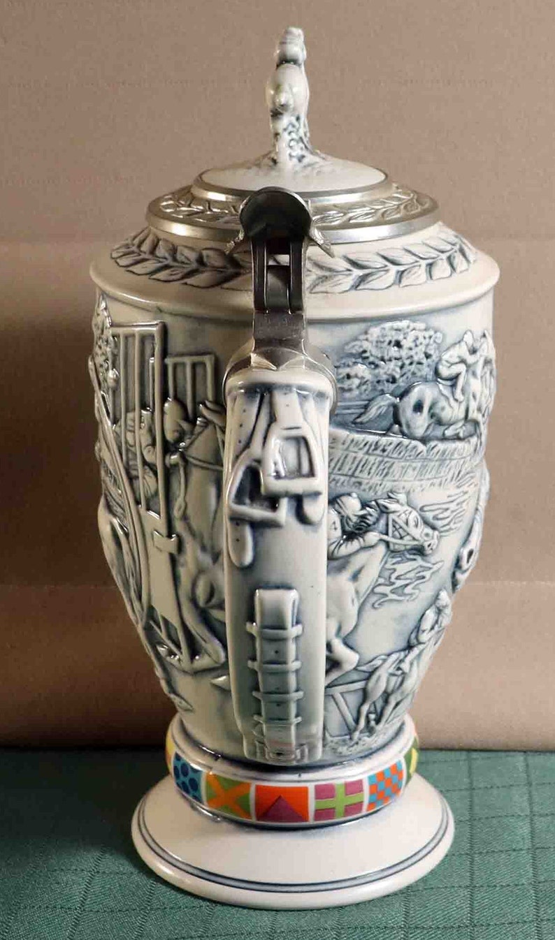 Puede incluir: Una jarra de cerveza de cer&aacute;mica con un dise&ntilde;o en relieve detallado que representa escenas ecuestres. La jarra tiene una tapa met&aacute;lica con bisagras, un asa decorativa y una banda colorida alrededor de la base. La combinaci&oacute;n de colores es blanca y azul.