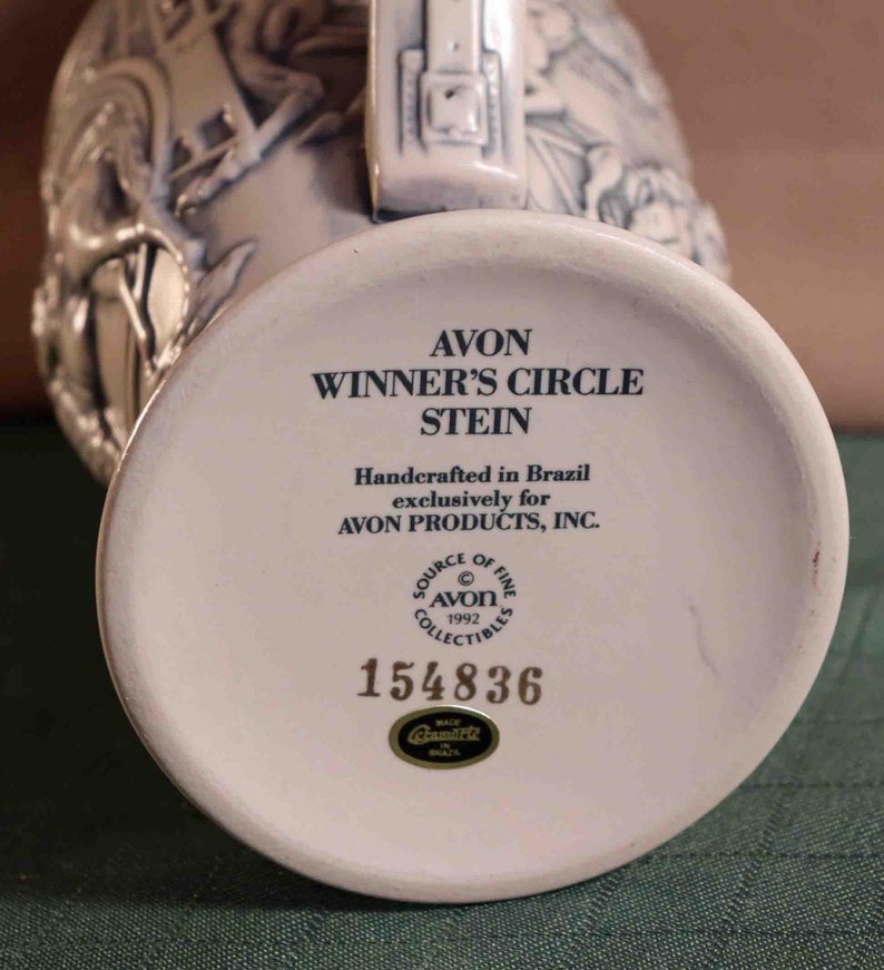 Puede incluir: Una jarra de cer&aacute;mica blanca con el texto "AVON WINNER'S CIRCLE STEIN" y "Handcrafted in Brazil". La base de la jarra tiene el n&uacute;mero "154836" y el logotipo de Avon. La jarra es un art&iacute;culo de colecci&oacute;n.