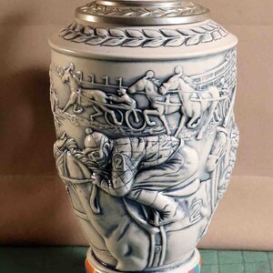 Puede incluir: Una jarra de cerveza de cer&aacute;mica blanca y azul con un relieve detallado de una escena de carreras de caballos. La tapa es plateada con una figura de caballo en la parte superior. La base presenta un patr&oacute;n geom&eacute;trico colorido.