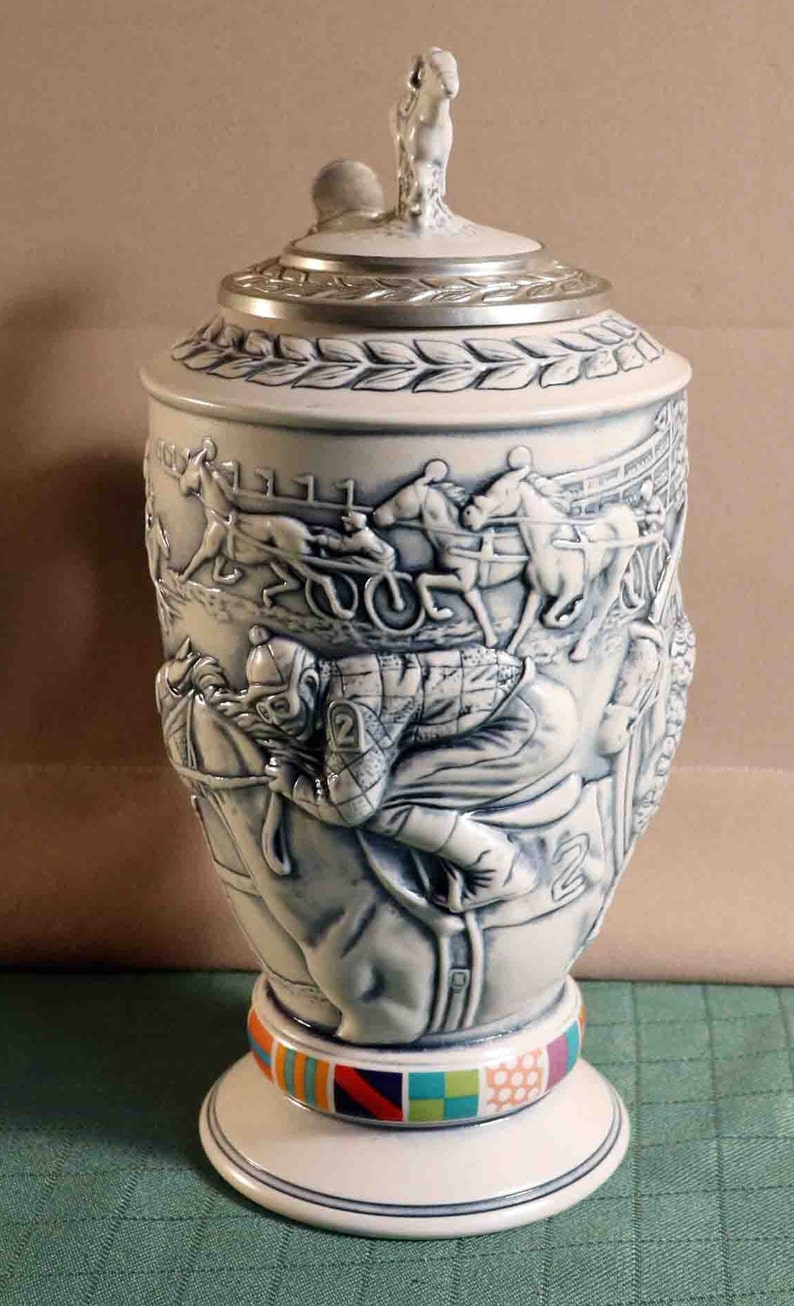 Puede incluir: Una jarra de cerveza de cer&aacute;mica blanca y azul con un relieve detallado de una escena de carreras de caballos. La tapa es plateada con una figura de caballo en la parte superior. La base presenta un patr&oacute;n geom&eacute;trico colorido.
