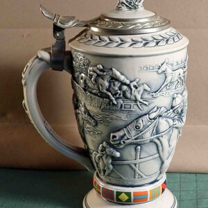 Puede incluir: Una jarra de cerveza de cer&aacute;mica con un relieve detallado de una escena de carreras de caballos. La tapa es de metal y tiene una peque&ntilde;a figura de caballo y jinete en la parte superior. El asa es curva y la base tiene un colorido patr&oacute;n geom&eacute;trico. La jarra mide unos 25 cm de altura.