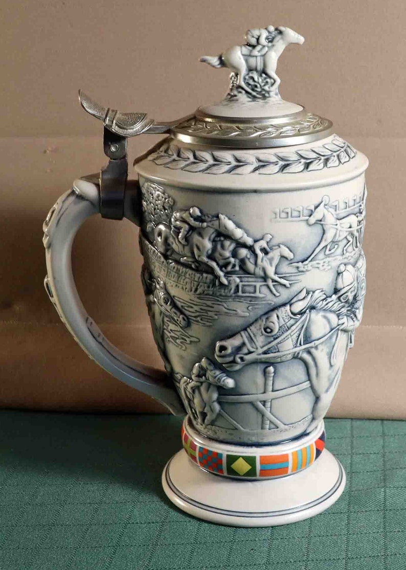 Puede incluir: Una jarra de cerveza de cer&aacute;mica con un relieve detallado de una escena de carreras de caballos. La tapa es de metal y tiene una peque&ntilde;a figura de caballo y jinete en la parte superior. El asa es curva y la base tiene un colorido patr&oacute;n geom&eacute;trico. La jarra mide unos 25 cm de altura.
