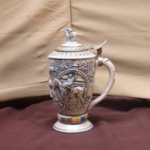 Puede incluir: Una jarra de cerveza de cer&aacute;mica con un relieve detallado de una escena de carreras de caballos. La jarra tiene una tapa con bisagras con una figura de caballo y jinete. La base presenta una banda colorida de cuadrados.