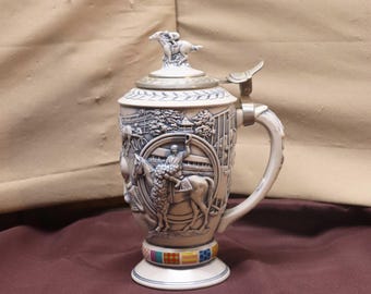 May include: A ceramic beer stein with a detailed relief of a horse race scene. The stein has a hinged lid with a horse and rider figurine. The base features a colorful band of squares. The handle and lid are silver-toned.