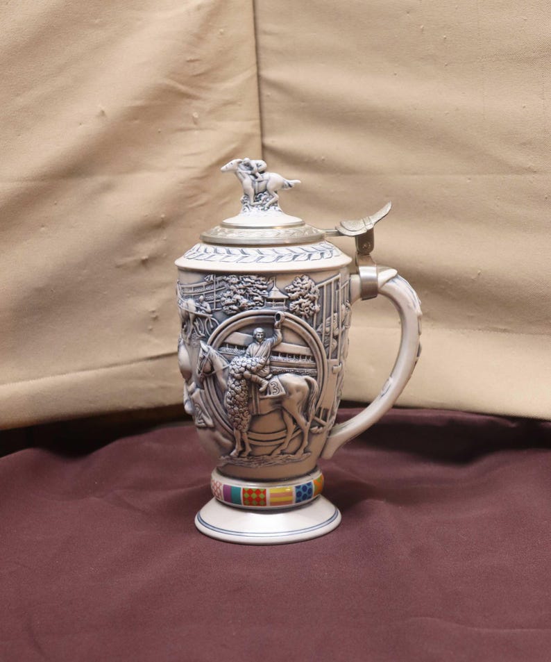 Puede incluir: Una jarra de cerveza de cer&aacute;mica con un relieve detallado de una escena de carreras de caballos. La jarra tiene una tapa con bisagras con una figura de caballo y jinete. La base presenta una banda colorida de cuadrados.
