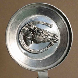 Puede incluir: Tapa de jarra de cerveza plateada con un relieve detallado de la cabeza de un caballo dentro de una herradura. El caballo lleva brida. La tapa tiene un dise&ntilde;o circular con un asa.