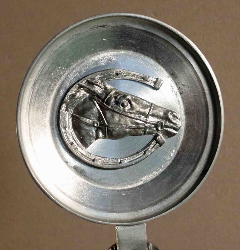 Puede incluir: Tapa de jarra de cerveza plateada con un relieve detallado de la cabeza de un caballo dentro de una herradura. El caballo lleva brida. La tapa tiene un dise&ntilde;o circular con un asa.
