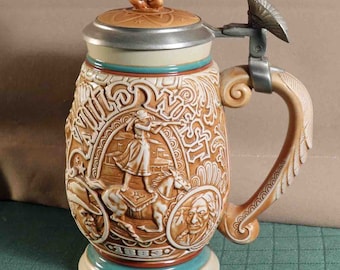 Puede incluir: Una jarra de cerveza de cerámica marrón con un diseño en relieve detallado que representa figuras y escenas históricas. La tapa presenta una pequeña figura nativa americana. El asa y la base tienen elementos decorativos.