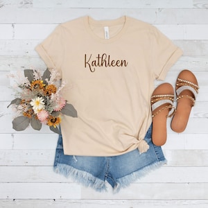 May include: A beige t-shirt with the name "Kathleen" printed in brown cursive lettering. The shirt is worn with denim shorts and brown sandals with rhinestones.