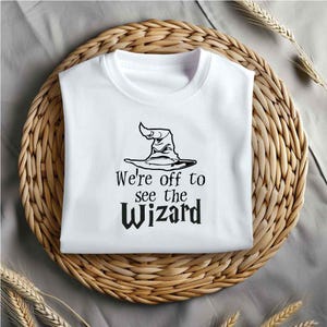 May include: White t-shirt with a black graphic of a wizard's hat and the text "We're off to see the wizard".