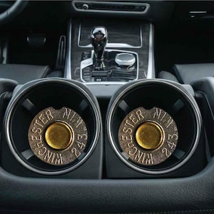 May include: Two circular cup holder inserts featuring a metallic finish with a textured base. Each insert displays the text Winchester 243 around a central gold-colored circular element, designed to fit into the console of a vehicle.