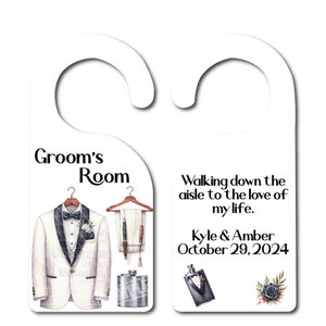 May include: White door hanger with a groom's room theme. The left side features a white tuxedo with a black bow tie and a pocket square. The right side has the text "Walking down the aisle to the love of my life. Kyle & Amber October 29, 2024".