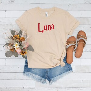 May include: A beige t-shirt with the word "Luna" printed in red on the front. The shirt is styled with denim shorts and sandals.