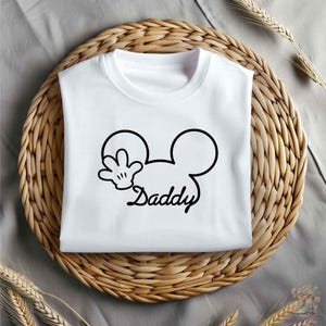 May include: White t-shirt with a black graphic of Mickey Mouse ears and hand, with the word "Daddy" in a script font. The shirt is folded and placed on a woven, circular placemat.