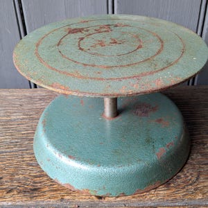 May include: A vintage green metal pottery wheel with a circular rotating top and a sturdy base. The surface features visible signs of wear, including rust patches and concentric circular markings on the top plate, showing a weathered texture.