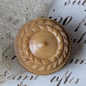 May include: A round, beige button featuring a tiered, concentric design with a central point and a textured, serrated outer edge. The button has a smooth, polished finish and exhibits subtle variations in its light tan color throughout its surface.