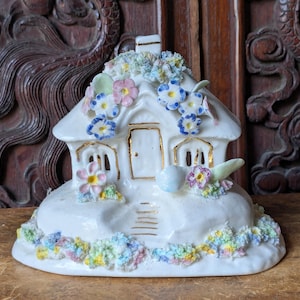 Könnte beinhalten: Eine weiße Keramik-Häuschen-Figur mit floralem Dach und Akzenten. Das Häuschen hat eine goldene Tür, Fenster und Verzierungen mit bunten Blumen und Blättern. Der Sockel ist ebenfalls mit bunten Blumen verziert.