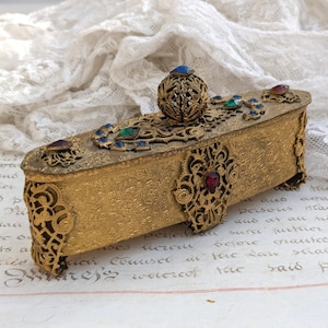 May include: A rectangular, antique gold-colored box with ornate filigree and jewel accents. The box features a hinged lid with a decorative ball and is adorned with red, blue, and green gemstones. The box is resting on a vintage paper with handwritten text.