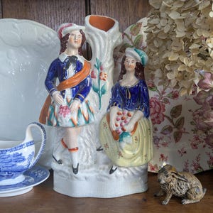Pode incluir: Uma figura de cerâmica vintage com duas figuras em trajes tradicionais, um homem e uma mulher, em pé ao lado de uma estrutura semelhante a uma árvore. As figuras estão vestidas de azul, laranja e branco. Uma molheira e um prato azuis e brancos estão em primeiro plano.