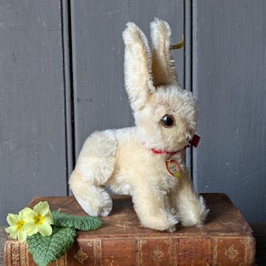 May include: A vintage plush rabbit toy with long ears, a red collar, and a small tag. The rabbit is cream-colored and sits on a brown book. Yellow flowers and green leaves are in the foreground. The background is a gray wooden surface.