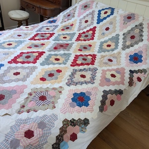Puede incluir: Una colcha de patchwork hecha a mano con un patrón hexagonal repetitivo. La colcha está compuesta por numerosos hexágonos de tela pequeños en varios colores y estampados, incluyendo rojo, azul, rosa y cuadros, unidos con bordes de tela blanca.