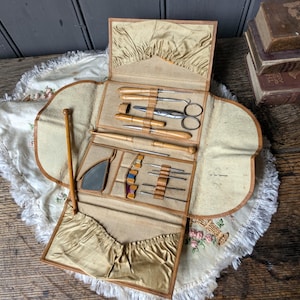 May include: A tan fabric sewing kit roll featuring a collection of metal and wooden tools including scissors, needles, and a small mirror. The interior is lined with beige satin pockets and elastic loops to secure each individual sewing implement.