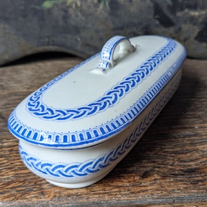 Peut inclure: Un plat ovale en céramique avec couvercle, base blanche et bordure tressée bleue. Le couvercle a une petite poignée et le plat repose sur une surface en bois. Le motif bleu est répété autour de la base du plat.