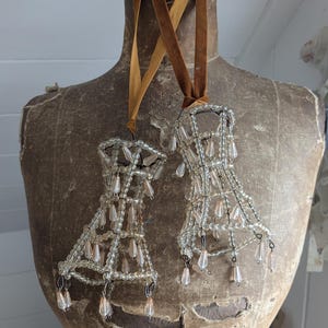 May include: Two decorative chandelier ornaments with clear beads and teardrop accents hang from brown velvet ribbons. The ornaments are displayed on a vintage mannequin torso, creating a rustic and elegant aesthetic.