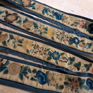 May include: A long, narrow fabric strip featuring a beige centre panel decorated with a repeating hand-painted floral pattern in shades of blue, brown, and green. The strip is bordered by dark blue fabric edges along the entire length of the piece.