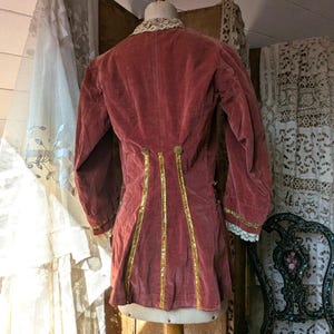 May include: A vintage, dusty rose velvet jacket with long sleeves and a lace collar. Gold trim accents the back and sleeves. The jacket is displayed on a mannequin, showcasing its antique design and details.
