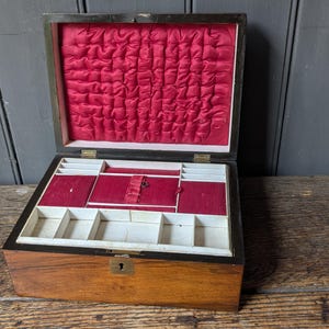 May include: A rectangular wooden jewelry box with a dark brown exterior and a brass keyhole. The interior lid features tufted red fabric, while the base contains multiple compartments lined with red and white fabric for organizing accessories.