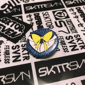 May include: A blue and yellow button with a cartoon image of a sailor collar with a yellow bow tie. The button has a white background with a blue and white polka dot pattern.