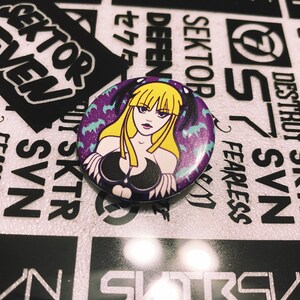 May include: A purple and black button featuring a cartoon image of a woman with blonde hair and a black outfit. The background is purple with a pattern of bats.