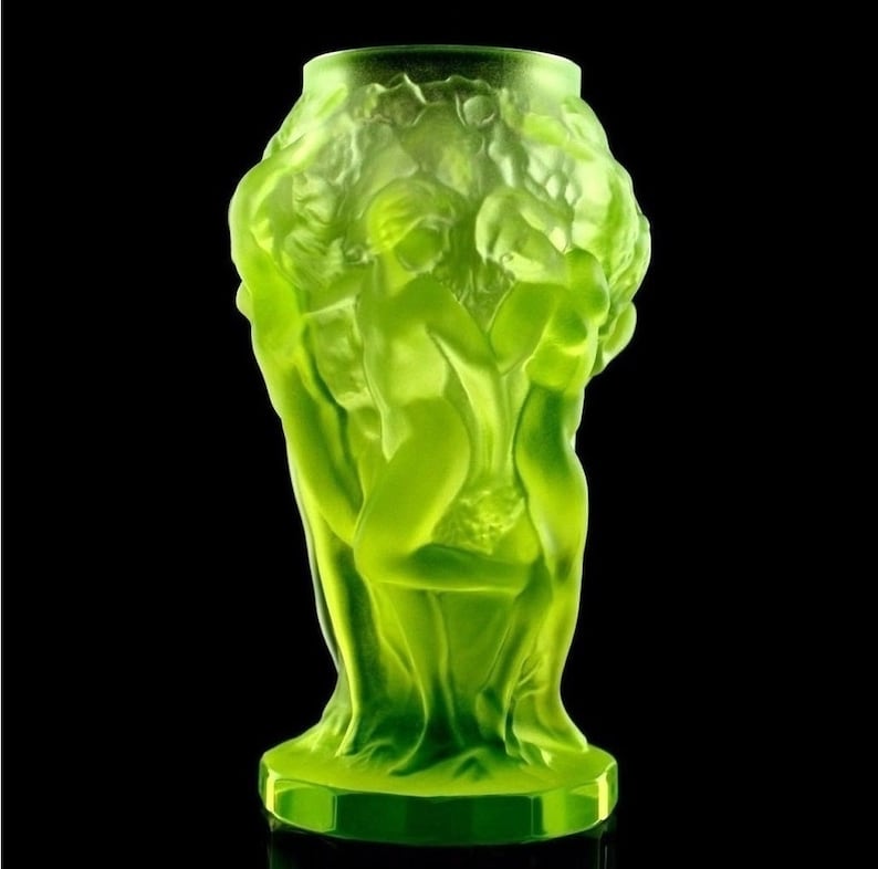 May include: A green glass vase with a sculpted design of three figures. The vase is made of frosted glass and has a rounded base.