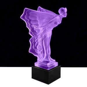 May include: A translucent purple glass sculpture depicting a figure with outstretched arms and large, wing-like drapery flowing behind. The figure is positioned in a forward-leaning pose and is mounted on a solid, square black base.
