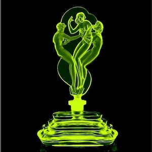 Peut inclure: Une sculpture de style art déco vert néon représentant trois figures féminines stylisées. La sculpture est montée sur une base à gradins et se détache sur un fond noir. Les figures sont en pose de danse.