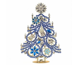 May include: A decorative Christmas tree ornament crafted from blue and clear rhinestones. The tree features a layered, swirling design accented with larger blue and clear crystal floral motifs, topped with a star-shaped crystal element.