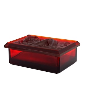 May include: A red glass box with a raised design of a reclining figure. The box has a clear lid and a dark red base.