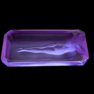 K&ouml;nnte beinhalten: Ein Aschenbecher aus violettem Glas mit einer geformten Figur einer liegenden Frau.