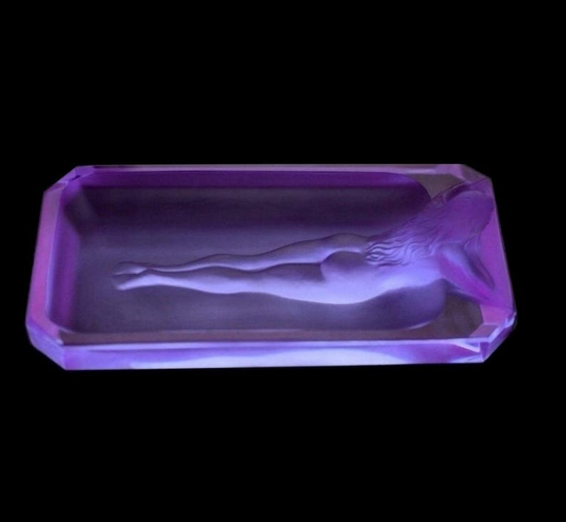 K&ouml;nnte beinhalten: Ein Aschenbecher aus violettem Glas mit einer geformten Figur einer liegenden Frau.