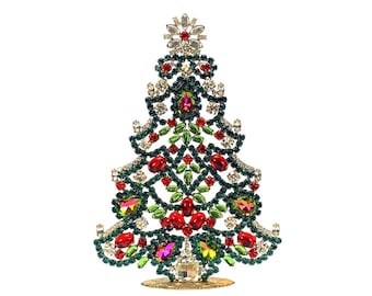 May include: A decorative Christmas tree ornament constructed from a metal frame encrusted with numerous small green rhinestones, larger red and green faceted jewels, and clear crystals, all arranged in a triangular, tiered, and branch-like pattern.