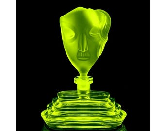 May include: A bright green glass bottle with a face-shaped stopper. The bottle has a layered base and is made of clear glass.