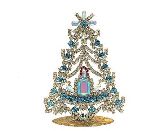 May include: A blue and white rhinestone Christmas tree ornament with a gold base. The tree is decorated with blue and clear rhinestones and a pink and blue iridescent center piece.