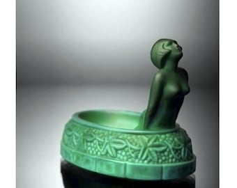 Puede incluir: Un cenicero de vidrio verde con una figura de mujer esculpida que emerge del lado. El cenicero está decorado con un patrón repetitivo de hojas y uvas.