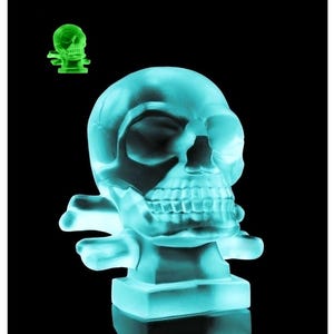 May include: A translucent, light blue skull sculpture resting on a square base with crossed bones behind it. A smaller, glowing green version of the same skull sculpture is positioned in the upper left corner against a solid black background.