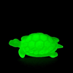 May include: A glowing green turtle figurine against a black background. The turtle has a textured shell and visible flippers, head, and tail. The bright green color suggests it may glow in the dark or be made of a luminescent material.