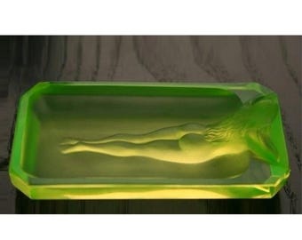 May include: A rectangular, translucent green ashtray with a sculpted nude figure reclining inside. The ashtray has a beveled edge and a smooth, polished surface. The figure is detailed with flowing hair and defined form.
