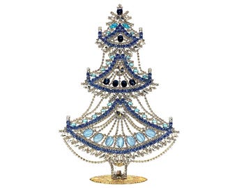 May include: A decorative Christmas tree sculpture with a gold base. The tree is adorned with silver-colored metal, blue and clear crystals, and oval-shaped blue stones. The tree's design features cascading chains and a star-like top.