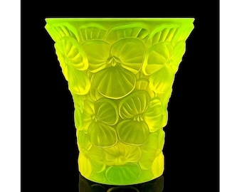 May include: A vivid, lime green vase featuring a detailed pansy floral pattern. The vase has a flared top and a cylindrical body, with the pansies intricately carved into the glass. The background is solid black, accentuating the vase's bright colour and design.